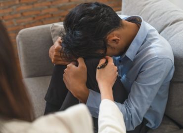 Stressed  Asian young man patient have life problem sitting on sofa with hands holding his head while woman psychiatrist trying to console him